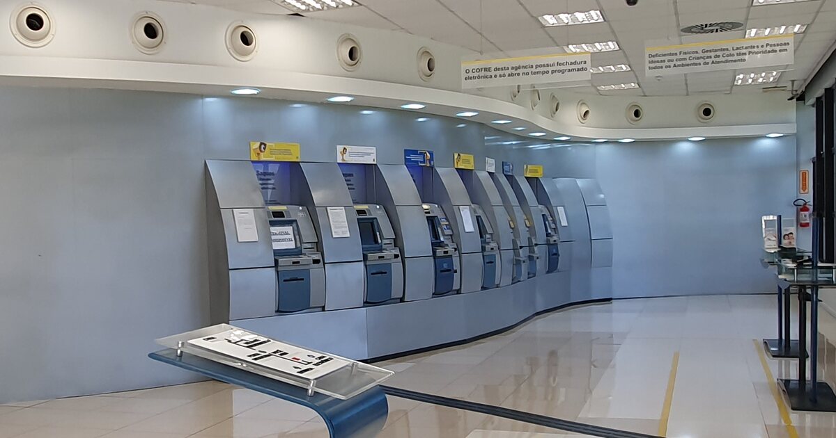 A curved row of seven ATMs in an empty Brazilian bank self-service hall, with Portuguese notices on the ceiling panel above them