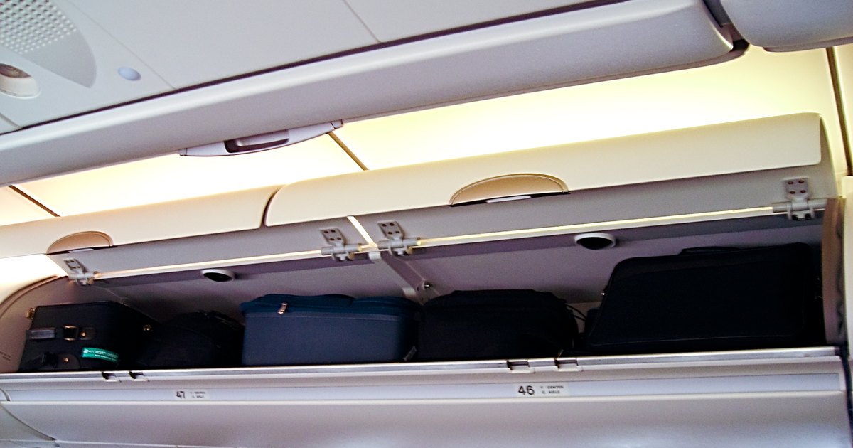 An open overhead locker on an airliner with five cabin cases packed side by side, the row markers 46 and 47 printed on the closed bin below