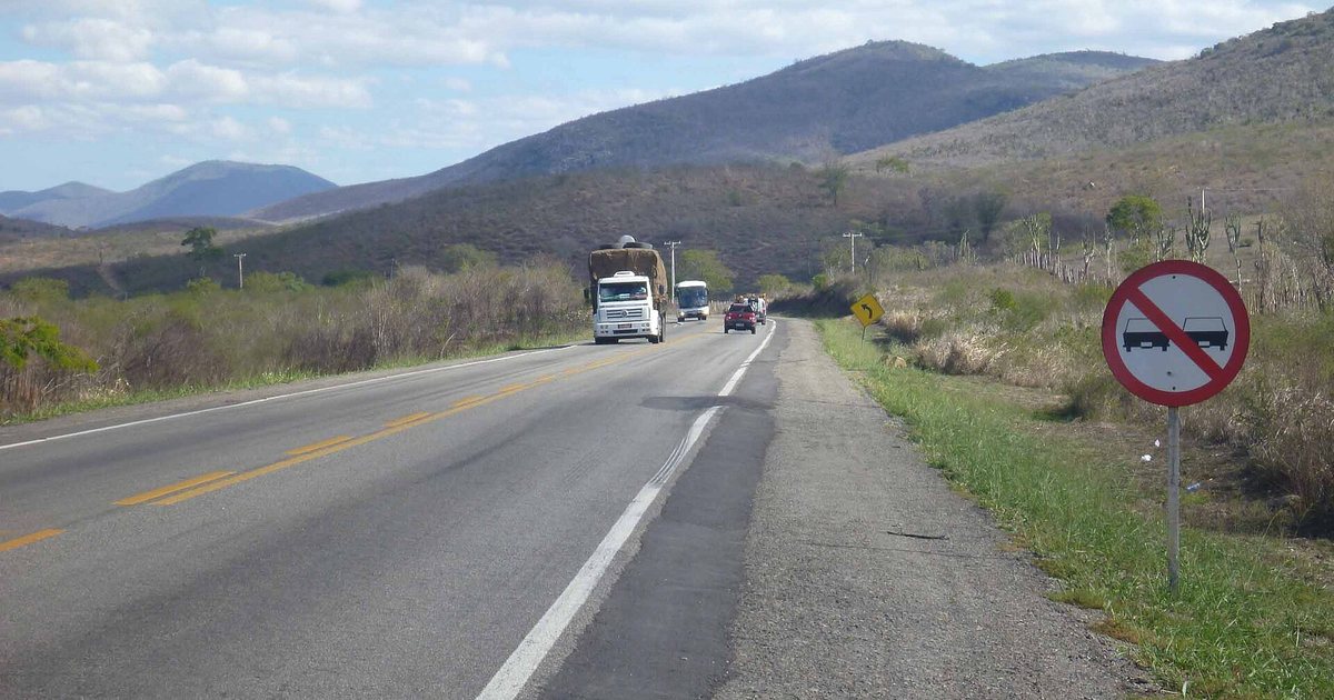 An undivided stretch of the BR-116 in Bahia with a double yellow centre line, a lorry, a bus and a car approaching in the opposite lane, and a no-overtaking sign