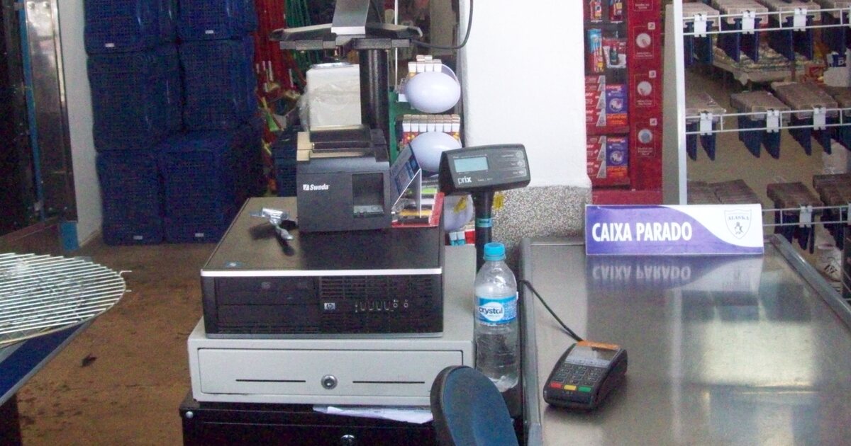 An empty supermarket checkout in Brazil: a fiscal receipt printer and cash drawer on the counter, a card pinpad on the steel belt, and a purple lane-closed sign