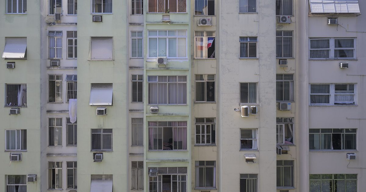 Facade of a tall Rio de Janeiro apartment block filling the frame, rows of near-identical windows with security grilles, roller shutters, window air conditioners and laundry hung out to dry