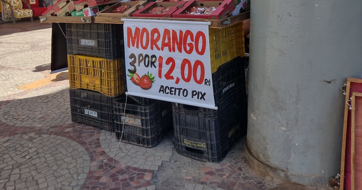 A strawberry stall on a Brazilian pavement, a printed banner across the stacked crates reading MORANGO 3 POR 12,00 R$ - ACEITO PIX