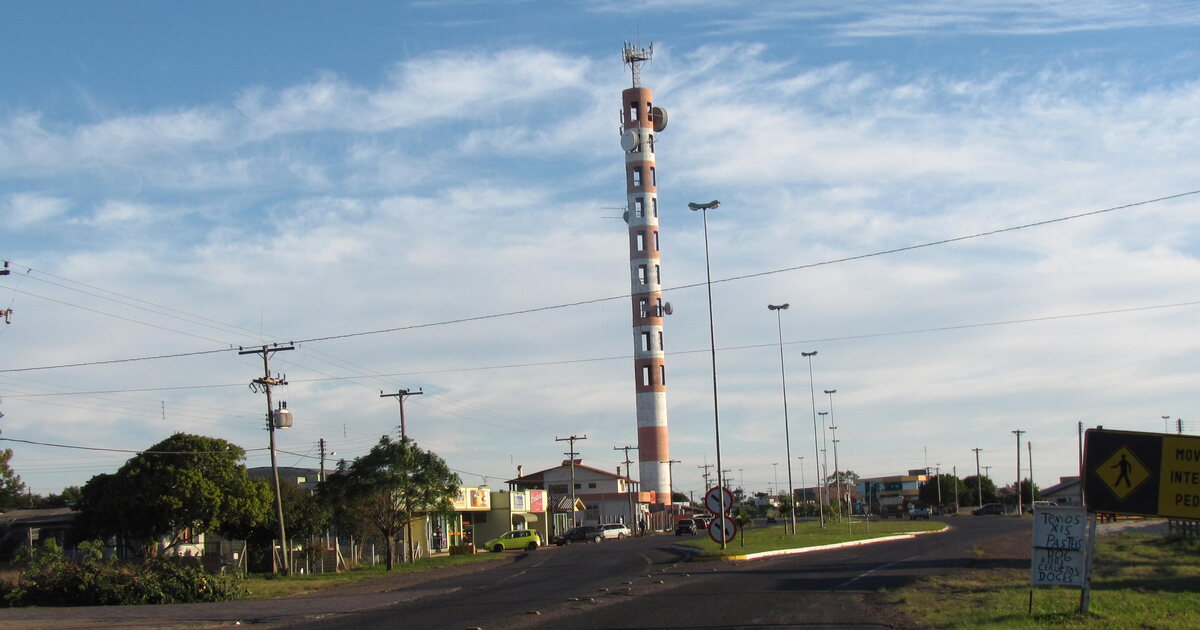A red and white mobile phone tower rising over the main road of a small Brazilian town, power poles and low shopfronts along the street