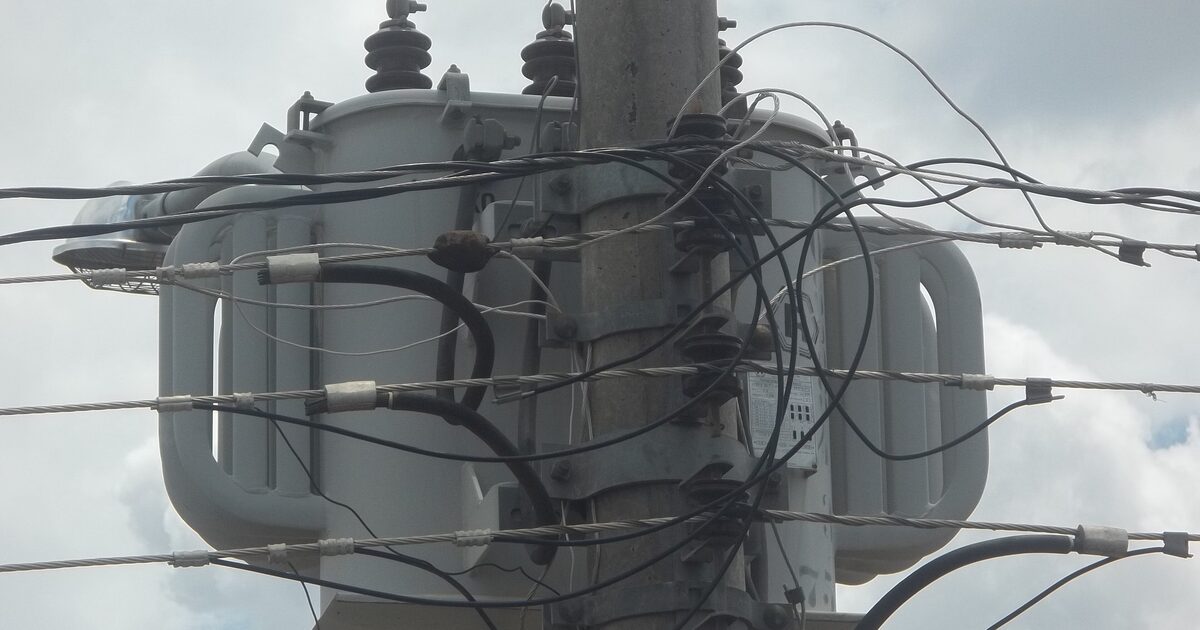 A pole-mounted electricity distribution transformer seen from below against a cloudy sky, low-voltage cables crossing in front of it